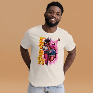 Puede incluir: Camiseta color crema con un gráfico colorido de un personaje femenino de estilo anime con cuernos, pantalones cortos y botas. El texto "PLAY SHAWN" está impreso verticalmente en amarillo y naranja.