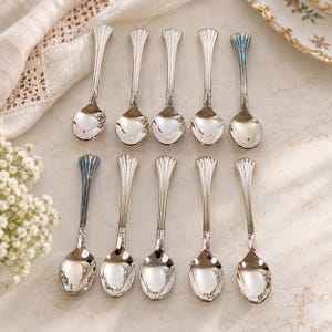 May include: A set of ten silver-toned metal spoons arranged in two rows of five. Each spoon features a slender handle with vertical fluted detailing and a polished, reflective bowl. The spoons have a uniform design with a flared top edge.