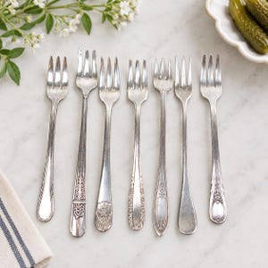 May include: A set of seven silver-toned metal cocktail forks arranged in a row. Each fork features a distinct handle design, including ornate floral engravings, geometric patterns, and smooth, plain finishes, all with three slender tines.