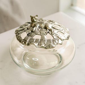 May include: A clear glass jar with a decorative lid. The lid is silver-coloured and features a cat figurine resting on a woven design. The jar has a round shape and is empty.
