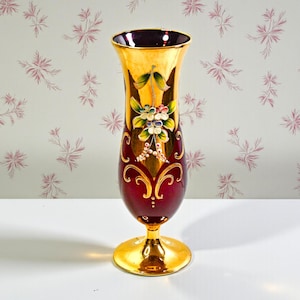 May include: A decorative vase with a gold rim and base, featuring a deep red body with gold accents and floral designs. The vase has a slender, elongated shape and is likely made of glass.