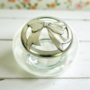 May include: A clear glass jar with a silver-coloured bow-shaped lid. The lid features a detailed bow design within a circular frame. The jar appears to be empty, with a slightly iridescent quality to the glass.