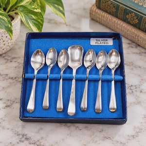 May include: A set of seven silver-plated spoons arranged in a blue velvet-lined box. The collection includes six oval-shaped spoons and one wider, square-edged spoon. A white label on the blue interior reads SILVER PLATED in black text.