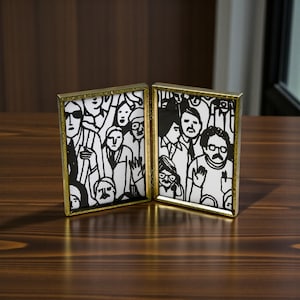 May include: Two-panel gold-framed artwork with black and white illustrations of people. The artwork is displayed on a dark wooden surface. The illustrations depict various faces and figures in a crowd.