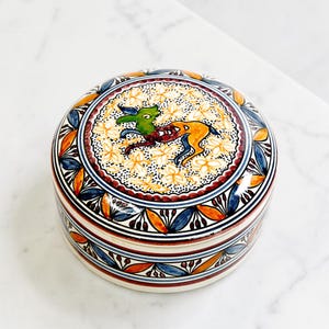 May include: A round, ceramic trinket box with a lid. The lid features a central design of a green and orange animal surrounded by yellow flowers. The box has a white base with blue, orange, and red floral patterns.