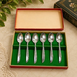 May include: A set of six silver-toned metal spoons arranged in a rectangular red box with a green velvet-like insert. Each spoon features a slender handle with a subtle embossed floral pattern along the stem and a smooth, polished oval bowl.
