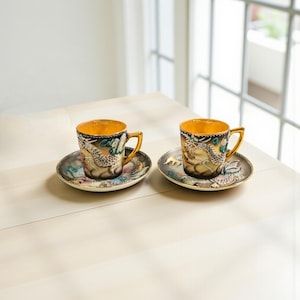 May include: Two ornate teacups and saucers. The cups have gold interiors and handles, with detailed dragon designs in shades of gray, teal, and pink. The saucers match the cups, creating a cohesive set. The set is placed on a light-colored surface.