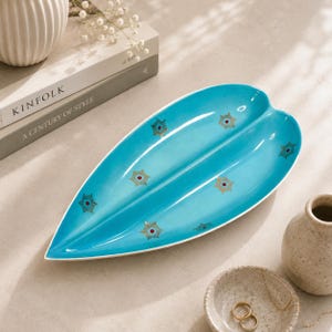 May include: A turquoise leaf-shaped ceramic serving dish with gold and red starburst designs. The dish has a white rim and a divided centre. Decorative home decor item.