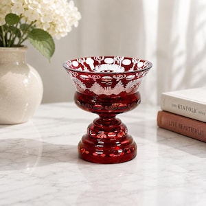May include: A deep red glass bowl with a pedestal base, featuring intricate white etched patterns around the rim and circular cut-out details along the body. The glass has a polished finish and a wide, flared opening at the top.
