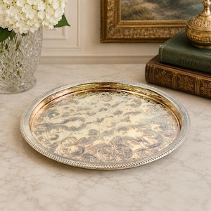 May include: A round silver-toned metal tray featuring a polished surface with an intricate, embossed scrollwork pattern and a beaded edge. The tray has a reflective finish that shows subtle gold-toned highlights across the central design.