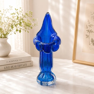 May include: A striking cobalt blue glass vase with a distinctive flared top and a textured, ribbed body. The vase features a rounded base and a clear, faceted bottom. The vase is empty and stands on a white surface.