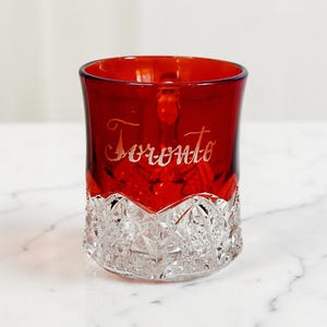 May include: A red glass tumbler with a clear, cut-glass base and the word "Toronto" etched on the side. The upper portion of the glass is a deep red, while the base features a diamond-cut pattern.