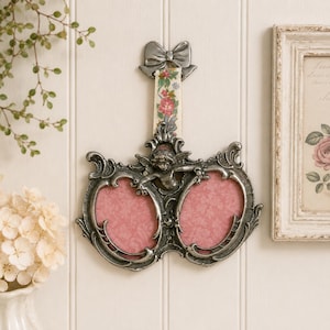 May include: Ornate, silver-toned double photo frame with an angel figurine and bow detail. The frame has two oval openings with pink backgrounds. A floral ribbon hangs from the top. Decorative wall art.