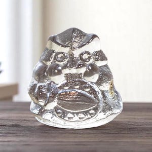 May include: A clear glass sculpture of a stylized face with prominent features, including large eyes and a wide mouth. The sculpture has a textured surface and sits on a wooden surface. The design is reminiscent of a mask or a decorative object.