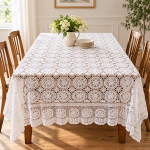 May include: A white rectangular tablecloth featuring an intricate crochet pattern of repeating circular motifs and a scalloped edge. The tablecloth is draped over a table, with a white ceramic pitcher of flowers and a stack of plates on top.