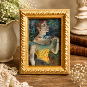 May include: A framed pastel painting of a woman in a yellow and blue dress. The painting is in a gold ornate frame. The artwork is displayed on a wooden surface, with a vase of flowers and books in the background.