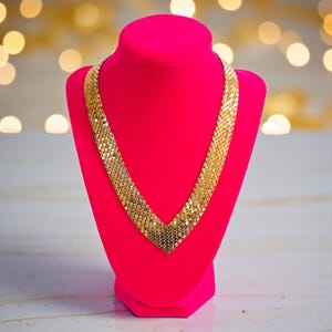 May include: A gold-coloured necklace with a V-shaped design is displayed on a bright pink jewellery bust. The necklace is made of small, interconnected rectangular pieces. The background features soft, blurred golden lights.