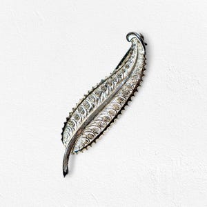 May include: A silver-toned leaf-shaped brooch with intricate filigree detailing. The brooch has a textured edge and a curved design, creating a delicate and elegant accessory. The brooch is set against a white background.