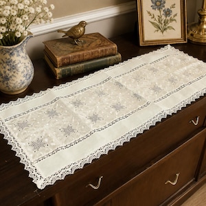 May include: Cream-colored table runner with a delicate star pattern and a scalloped lace edge. The runner is placed on a dark wooden dresser, with a vase of white flowers and antique books in the background.