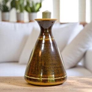 May include: A brown ceramic vase with a narrow neck and a wide base. The vase has a glossy finish with white and yellow stripes. A small, cylindrical stopper sits on top. The vase is on a wooden surface.