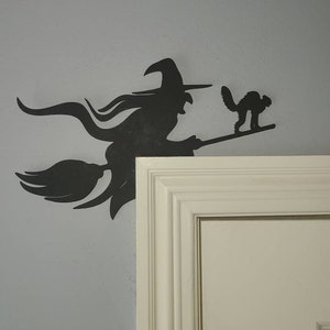 May include: Black silhouette of a witch flying on a broomstick with a black cat beside her. The silhouette is mounted on a white wall next to a white door.