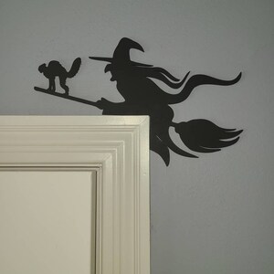 May include: Black silhouette of a witch flying on a broomstick with a black cat beside her. The silhouette is mounted on a white door frame.