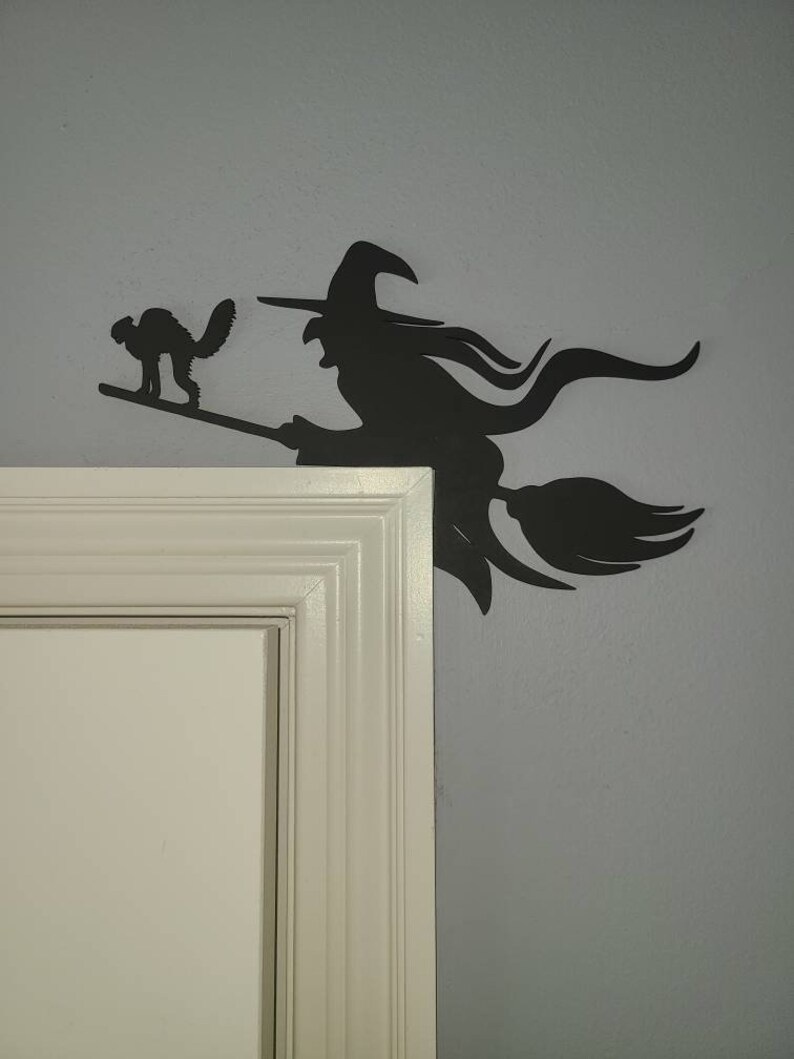 May include: Black silhouette of a witch flying on a broomstick with a black cat beside her. The silhouette is mounted on a white door frame.