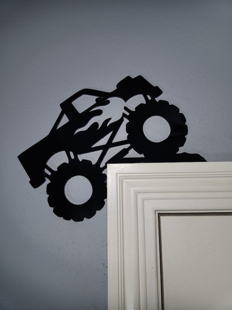 May include: Black silhouette of a monster truck with flames on the side, driving up a wall. The truck has large tires and is partially obscured by a white door frame.
