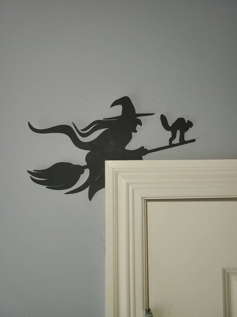 May include: Black silhouette of a witch flying on a broomstick with a black cat beside her. The silhouette is mounted on a white wall next to a white door.
