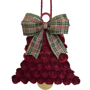 Puede incluir: Una decoración de pared con forma de campana cubierta de rosas de papel de color rojo oscuro. La parte superior presenta un gran lazo a cuadros en tonos verde, rojo y dorado. Un pequeño círculo de purpurina dorada está fijado en el centro inferior.