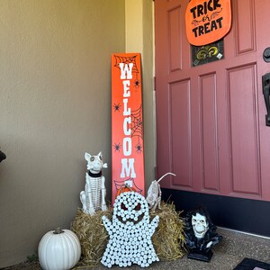 Puede incluir: Un letrero vertical alto de color naranja con la palabra WELCOME en letras blancas, decorado con telara&ntilde;as y ara&ntilde;as negras. Un letrero en forma de calabaza cuelga de la puerta con el texto TRICK or TREAT. Incluye una calabaza blanca y esqueletos.