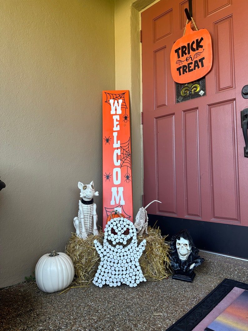 Puede incluir: Un letrero vertical alto de color naranja con la palabra WELCOME en letras blancas, decorado con telara&ntilde;as y ara&ntilde;as negras. Un letrero en forma de calabaza cuelga de la puerta con el texto TRICK or TREAT. Incluye una calabaza blanca y esqueletos.