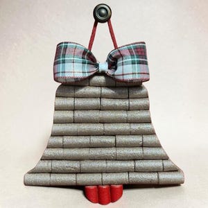 Puede incluir: Un adorno en forma de campana construido con filas horizontales de corchos texturizados de color marrón claro. La parte superior presenta un lazo de cinta a cuadros en tonos rojo, verde y blanco, unido a un aro de metal oscuro. Una pieza de madera roja cuelga en la base.