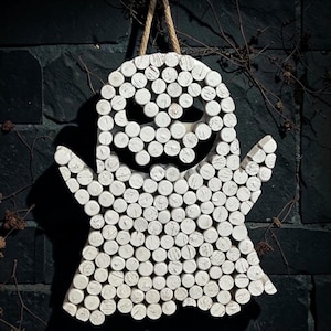 Puede incluir: Un adorno de pared con forma de fantasma, construido con numerosos corchos cil&iacute;ndricos pintados de blanco dispuestos para formar una figura con dos brazos y una boca dentada. Un lazo de cuerda color canela est&aacute; unido a la parte superior para colgar.