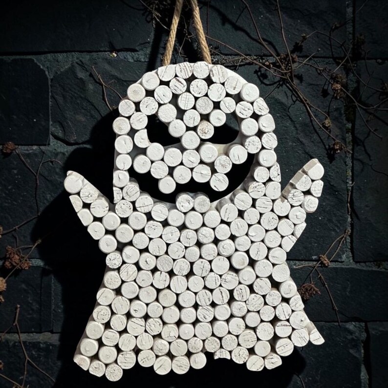 Puede incluir: Un adorno de pared con forma de fantasma, construido con numerosos corchos cil&iacute;ndricos pintados de blanco dispuestos para formar una figura con dos brazos y una boca dentada. Un lazo de cuerda color canela est&aacute; unido a la parte superior para colgar.