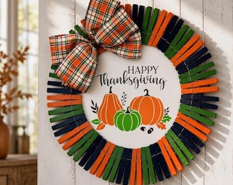 May include: A circular wreath constructed from alternating orange, green, and dark blue clothespins. A large plaid bow in orange, green, and white sits at the top. The center features a white disc with the text Happy Thanksgiving and pumpkins.