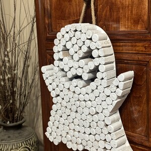 Puede incluir: Un adorno de pared con forma de fantasma construido con numerosas piezas cil&iacute;ndricas de madera pintadas de blanco y dispuestas en un patr&oacute;n denso. La figura presenta dos huecos circulares oscuros para los ojos y cuelga de una cuerda de yute.