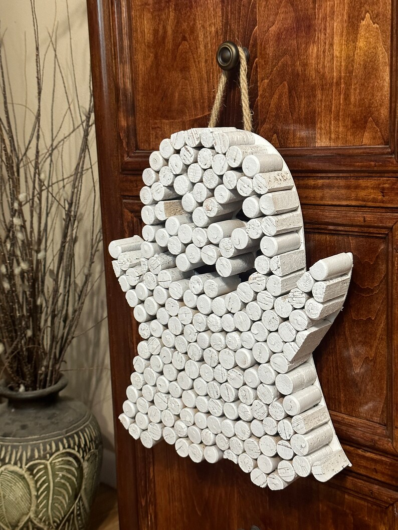 Puede incluir: Un adorno de pared con forma de fantasma construido con numerosas piezas cil&iacute;ndricas de madera pintadas de blanco y dispuestas en un patr&oacute;n denso. La figura presenta dos huecos circulares oscuros para los ojos y cuelga de una cuerda de yute.