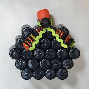 Puede incluir: Una decoración con forma de calabaza construida con múltiples tapas cilíndricas negras dispuestas en racimo. Presenta un tallo naranja brillante en la parte superior y una cinta decorativa con bordes ondulados verde lima y rayas de purpurina naranja.
