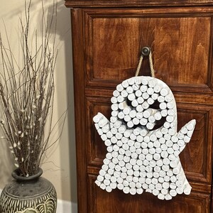 Puede incluir: Un adorno de pared con forma de fantasma hecho de numerosas piezas de madera cil&iacute;ndricas pintadas de blanco, dispuestas para formar una figura con dos brazos y aberturas oscuras para los ojos y la boca, colgado de una cuerda de yute.