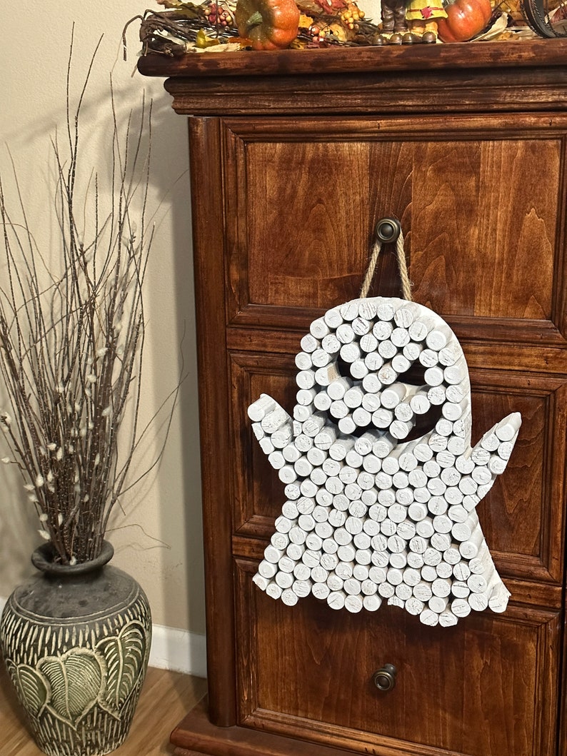 Puede incluir: Un adorno de pared con forma de fantasma hecho de numerosas piezas de madera cil&iacute;ndricas pintadas de blanco, dispuestas para formar una figura con dos brazos y aberturas oscuras para los ojos y la boca, colgado de una cuerda de yute.