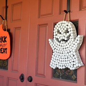 Puede incluir: Un letrero naranja con forma de calabaza con el texto negro "TRICK OR TREAT" y una decoraci&oacute;n con forma de fantasma hecha de numerosos corchos de vino blancos, ambos colgados de ganchos de metal negro en una puerta doble de color marr&oacute;n rojizo.