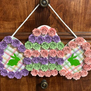 Puede incluir: Una corona de Pascua hecha a mano con tres paneles en forma de corazón decorados con rosetas de papel rosa, verde y morado. Cada panel presenta un fondo a cuadros y una silueta de conejo verde.