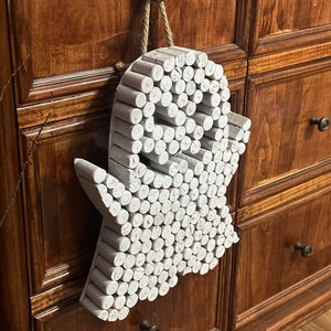 Puede incluir: Un adorno de pared con forma de fantasma construido con numerosas piezas de madera cil&iacute;ndricas pintadas de blanco dispuestas en un patr&oacute;n denso. El objeto presenta una cabeza redondeada, dos brazos extendidos y una base ondulada, colgado de un cordel de yute.
