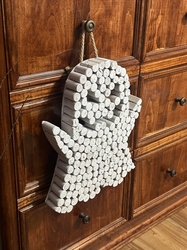 Puede incluir: Un adorno de pared con forma de fantasma construido con numerosas piezas de madera cil&iacute;ndricas pintadas de blanco dispuestas en un patr&oacute;n denso. El objeto presenta una cabeza redondeada, dos brazos extendidos y una base ondulada, colgado de un cordel de yute.