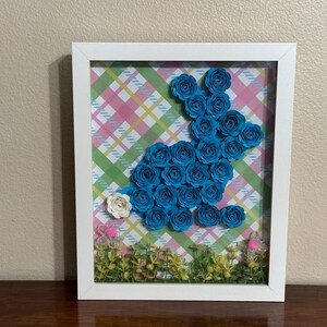 Puede incluir: Una artesanía de papel enmarcada de un conejito azul hecho de rosetas de papel. El conejito está sobre un fondo de cuadros rosa y verde con hierba verde y flores en la parte inferior.