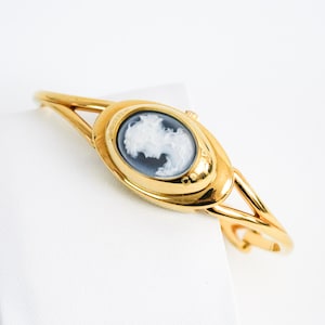 Puede incluir: Un reloj tipo brazalete dorado con una esfera ovalada que presenta un retrato de perfil de camafeo en blanco y negro. El reloj tiene un acabado de metal pulido, un diseño de banda dividida y una pequeña corona lateral. La palabra Cameo está grabada.
