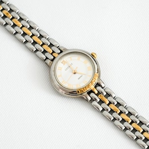 Puede incluir: Un reloj de pulsera Lucent Seiko plateado y dorado con esfera blanca y números romanos dorados. La correa del reloj es una combinación de eslabones plateados y dorados. La esfera del reloj dice "LUCENT" y "SEIKO".
