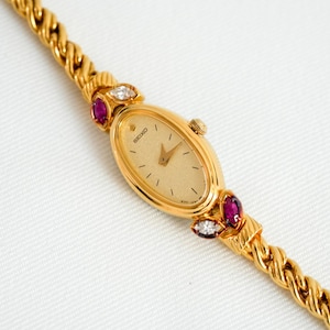 May include: A gold-toned Seiko wristwatch with an oval face and a gold band. The watch face is cream-colored with gold hands and markers. The band features ruby and diamond accents. The watch is on a white background.