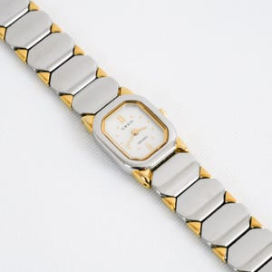May include: A Seiko Creo wristwatch featuring a rectangular white dial with gold-tone hands and markers. The watch has a two-tone link bracelet constructed from silver-tone rectangular segments connected by small gold-tone triangular accents.